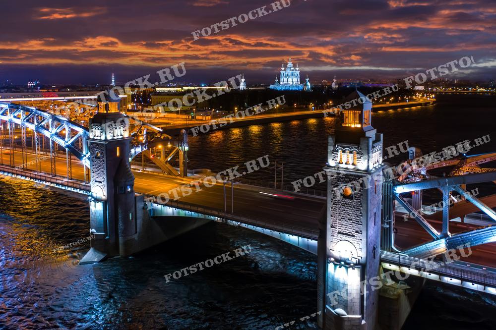 На фотографии изображен Большеохтинский мост в Санкт-Петербурге ночью. Мост ярко освещен, создавая контраст с темным небом. На заднем плане виден Смольный собор, также подсвеченный огнями. Вечернее небо с облаками придает картине дополнительную привлекательность.